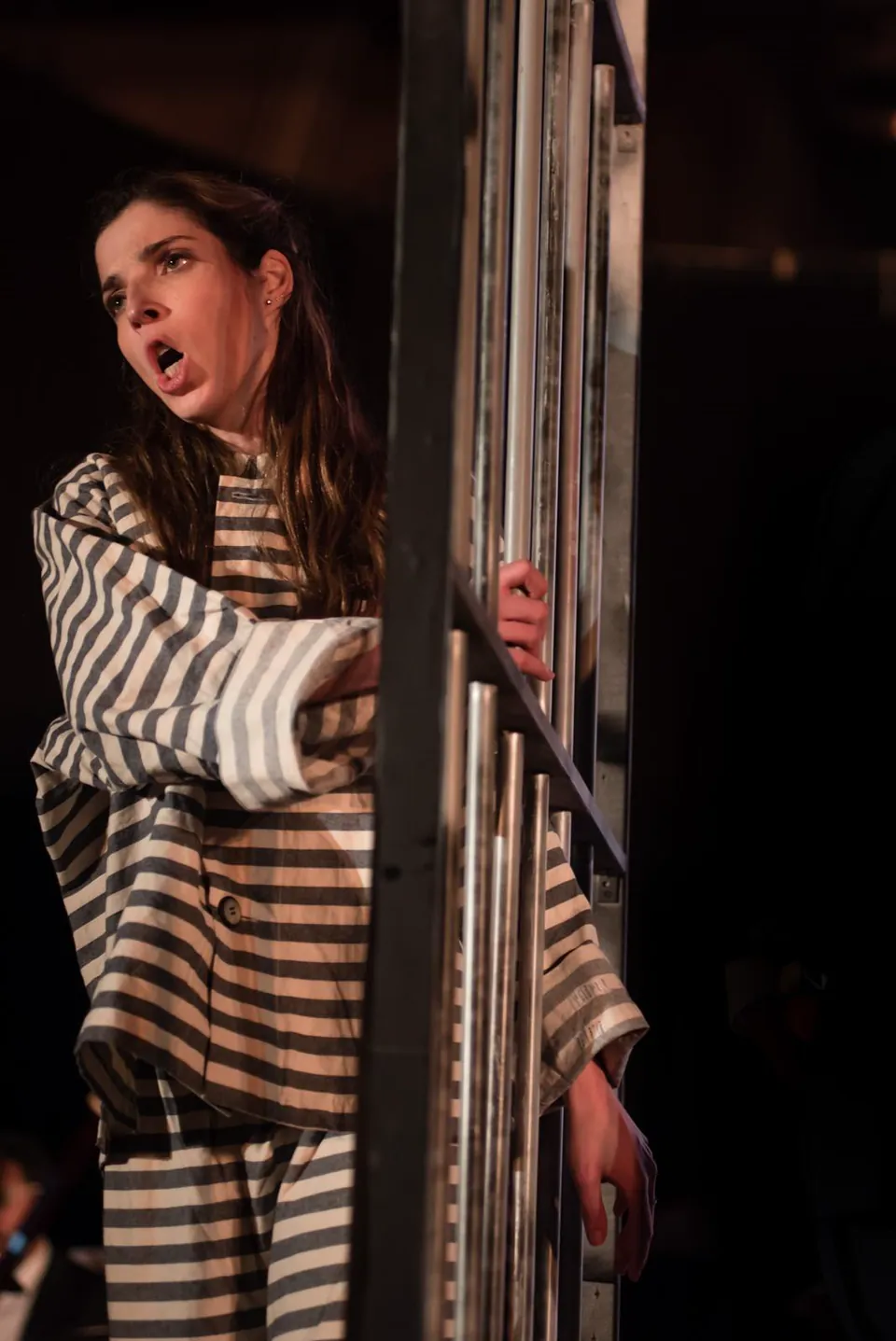Monica on stage in a striped costume, singing with her mouth open and one hand gripping metal bars.