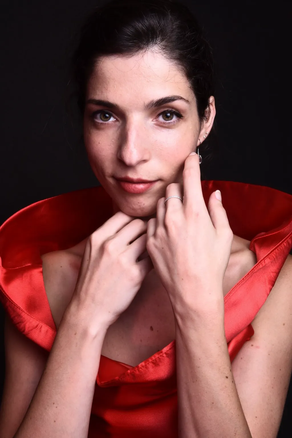 Portrait of Monica Schwartz in a red satin garment with a high collar, hands raised near her chin and cheek, looking at the camera against a dark background.